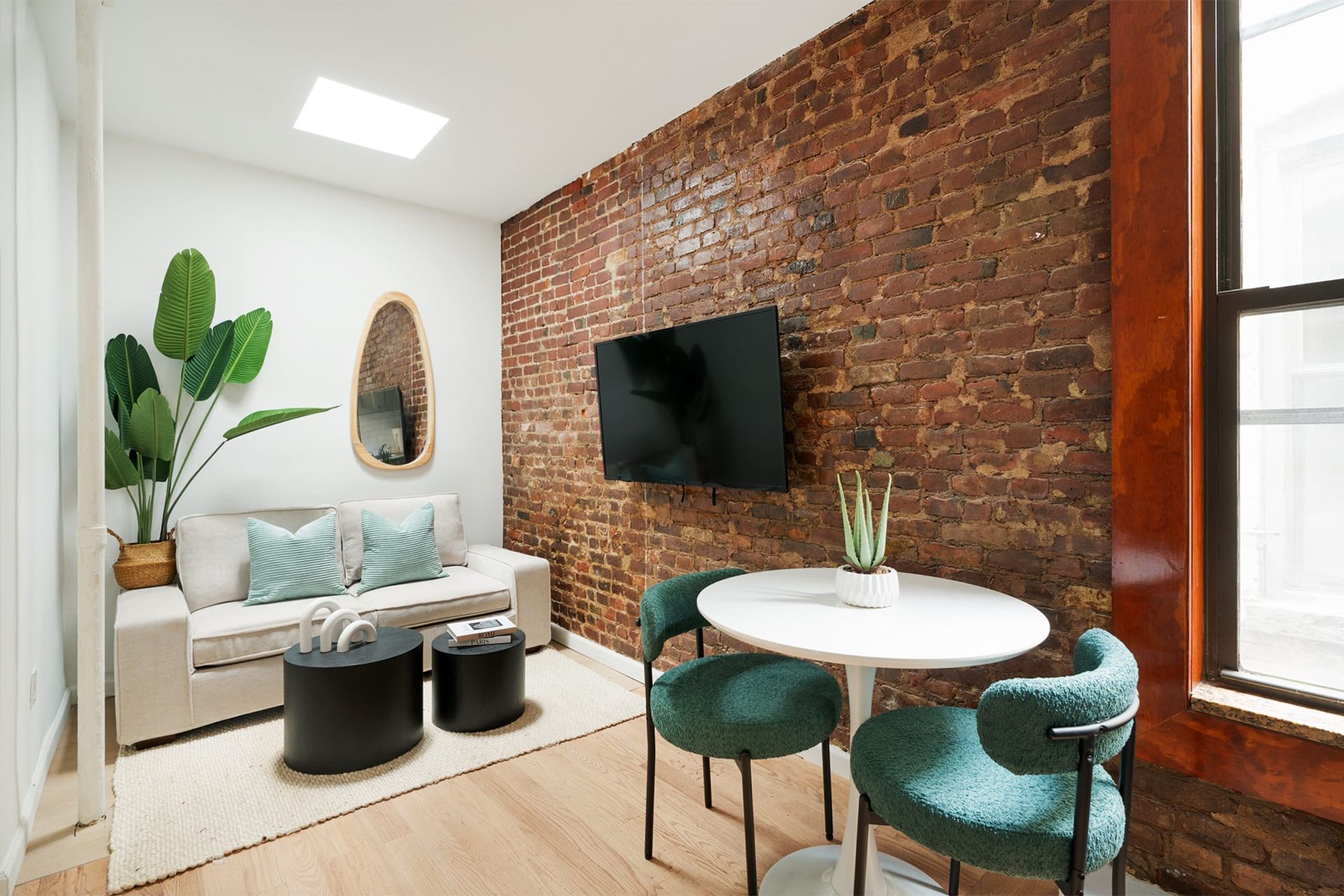 Bright 2-bedroom apartment in the Lower East Side with a washer and dryer, showcasing an exposed brick wall, a comfortable beige sofa with green accent pillows, a round white dining table with two green cushioned chairs, and a sleek black coffee table set. The space includes a flat-screen TV and is adorned with a decorative wall mirror and indoor plants.
