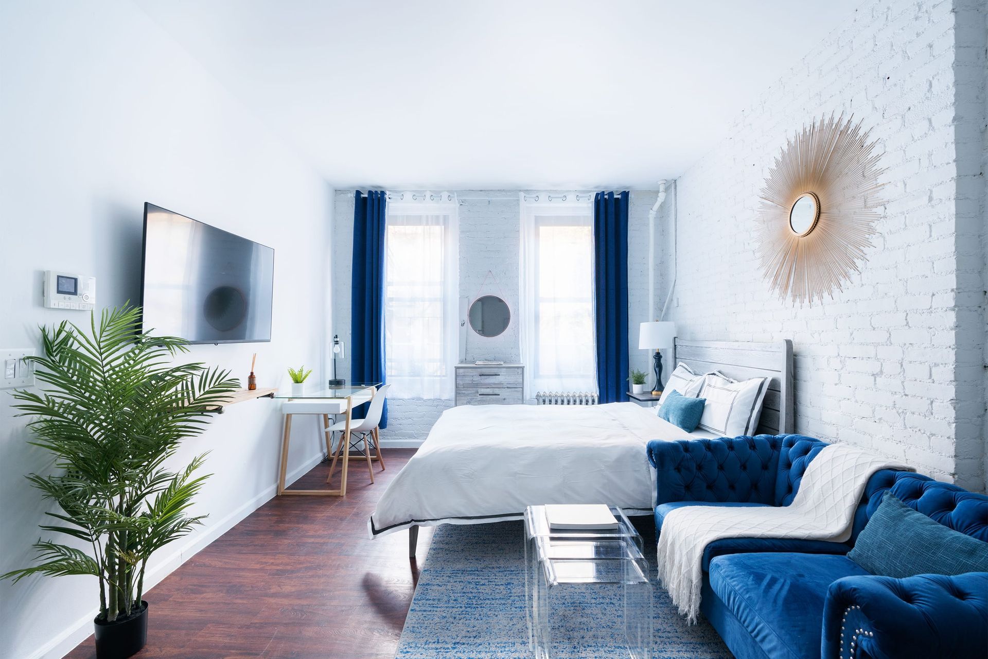Bright studio apartment with a white bed, blue armchair, clear coffee table, potted plant, wall-mounted TV, large windows with blue curtains, and a decorative sunburst mirror on a white brick wall. The apartment is located in the Upper East Side.