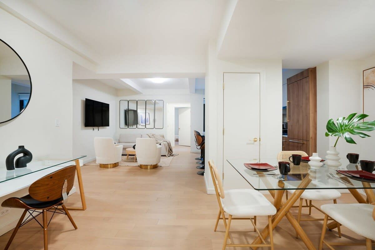 Spacious, bright apartment interior with dining and living areas. Light wood floors, white walls, modern furniture.