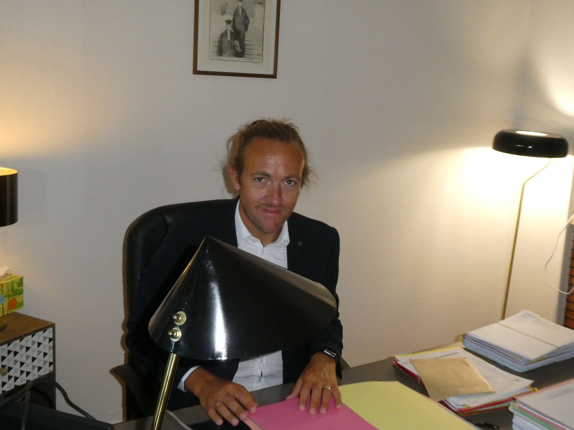 Une personne assise à un bureau dans un bureau, avec des papiers, une lampe et un écran d'ordinateur visibles.