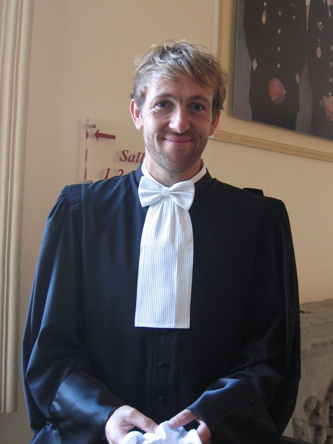Un homme vêtu d'une robe noire et d'une cravate blanche, debout à l'intérieur et souriant à la caméra.