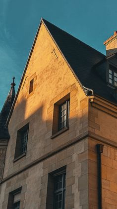 Bâtiment en pierre baigné de soleil, avec un toit sombre et pentu et de hautes fenêtres se détachant sur un ciel bleu.