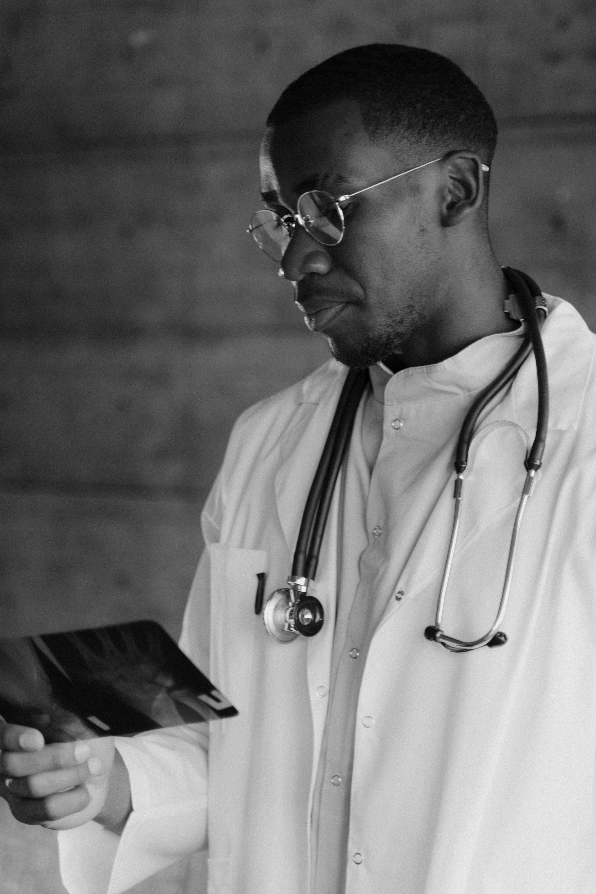 Un médecin en blouse blanche, stéthoscope à la main, consulte une tablette dans un décor gris.