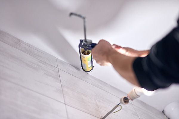 Eine Hand hält eine Sprühdose über einen grauen Fliesenboden, daneben liegt ein Pinsel auf dem Boden.