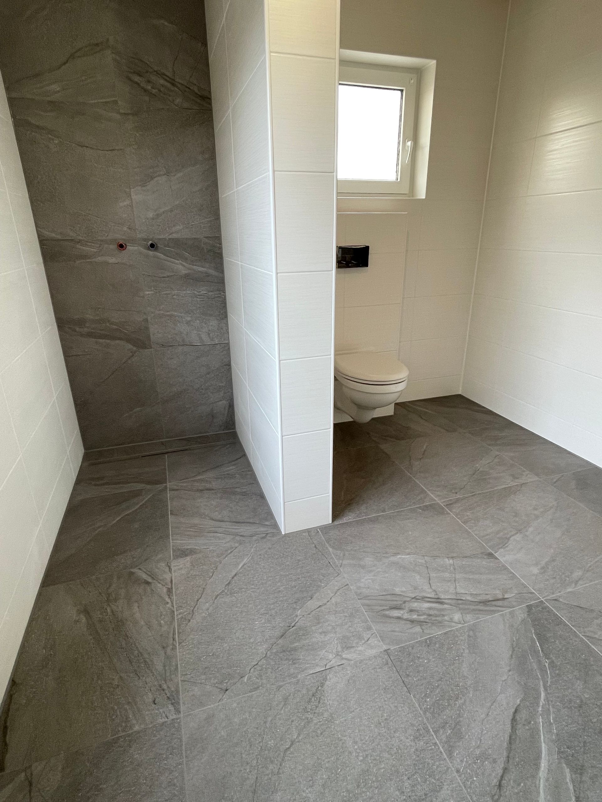 Unfertiges Badezimmer mit gefliestem Boden, teilweise abgehängter Wand, Fenster und sichtbarem Toilettenbereich