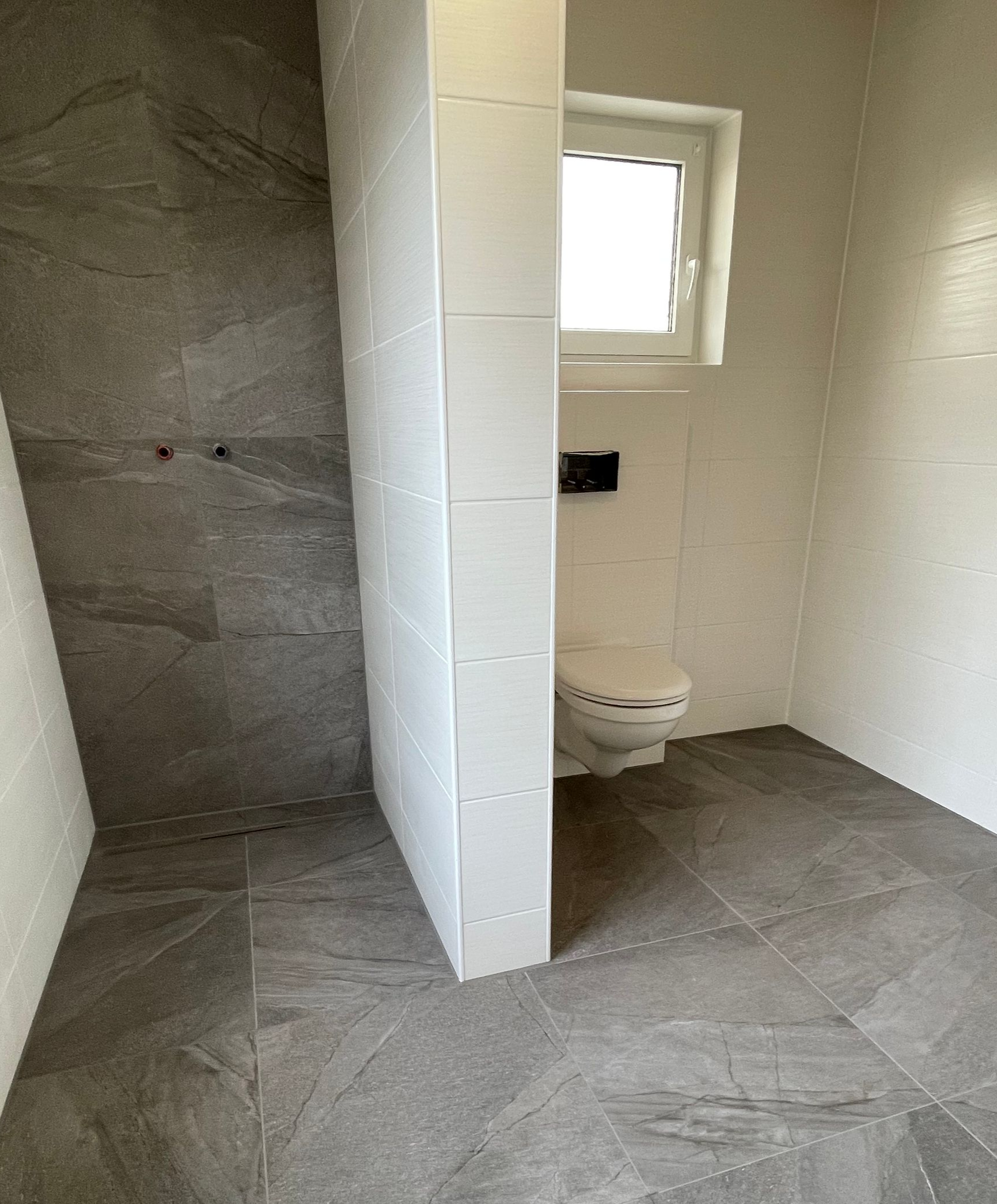 Kleines, gefliestes Badezimmer mit Toilette, Fenster und Duschbereich, vom Eingang aus einsehbar