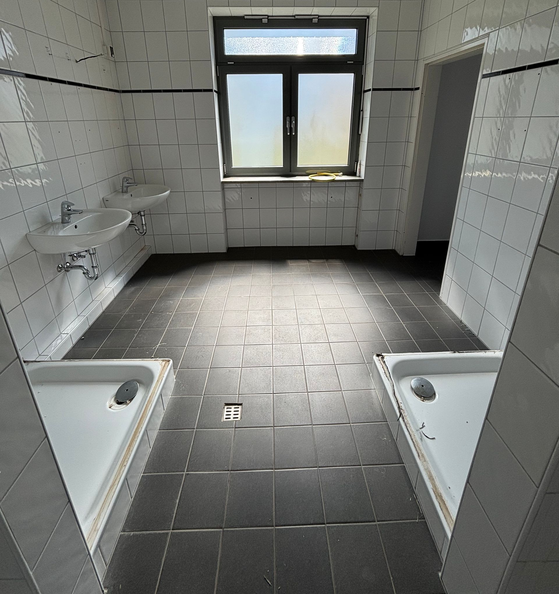 Leere, geflieste öffentliche Toilette mit zwei Duschen, einem wandmontierten Waschbecken und einem Fenster.