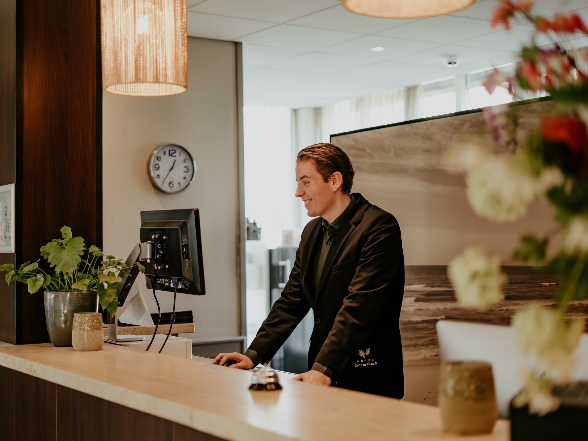 Een jonge man in een zwart jasje met logo van Hotel Heemskerk, staat lachend achter de receptiebalie van Hotel Heemskerk en kijkt naar een computerscherm.