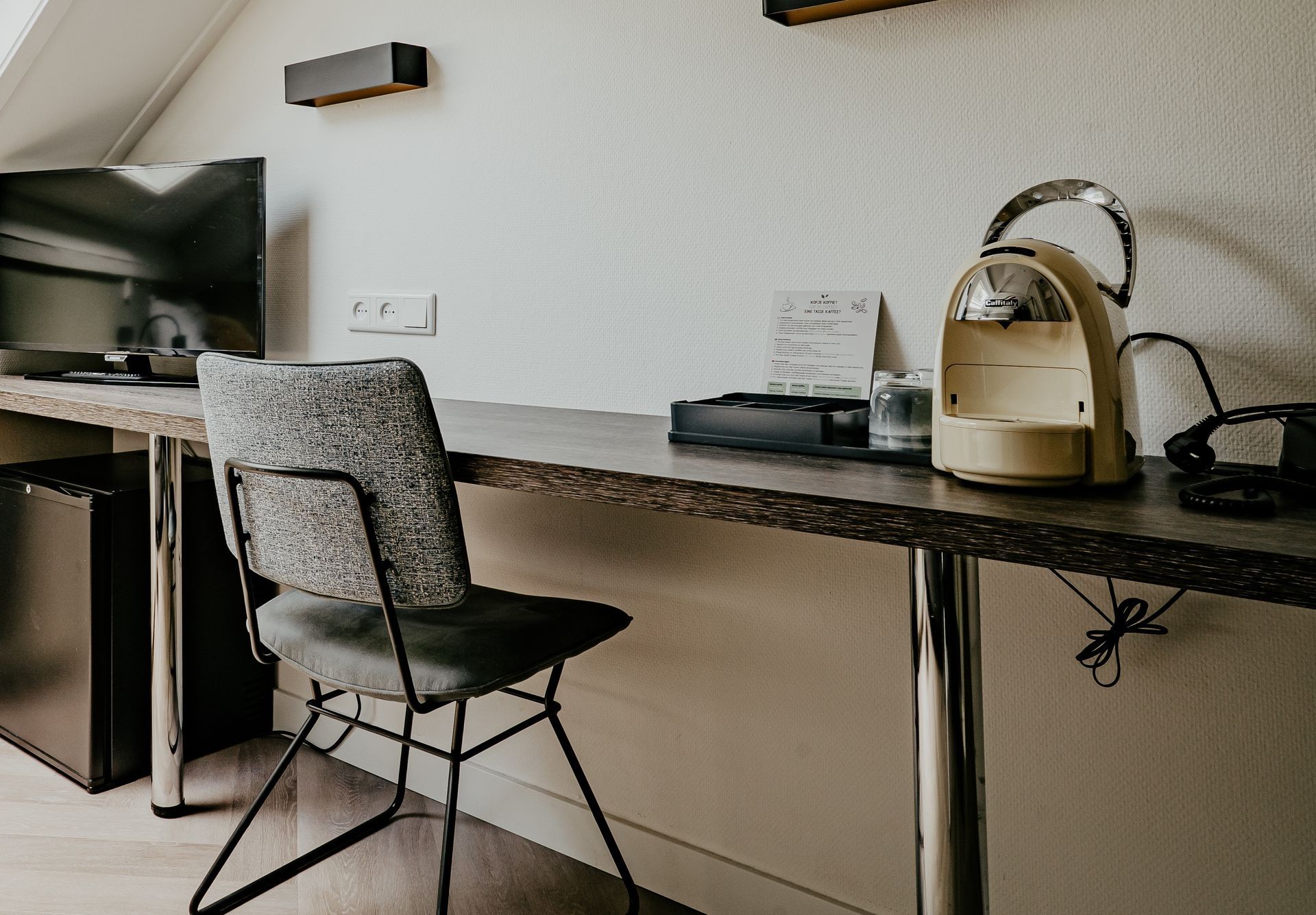 A desk in the single room at Hotel Heemskerk featuring a comfortable chair, flat-screen TV, coffee machine, glass, and guest information.