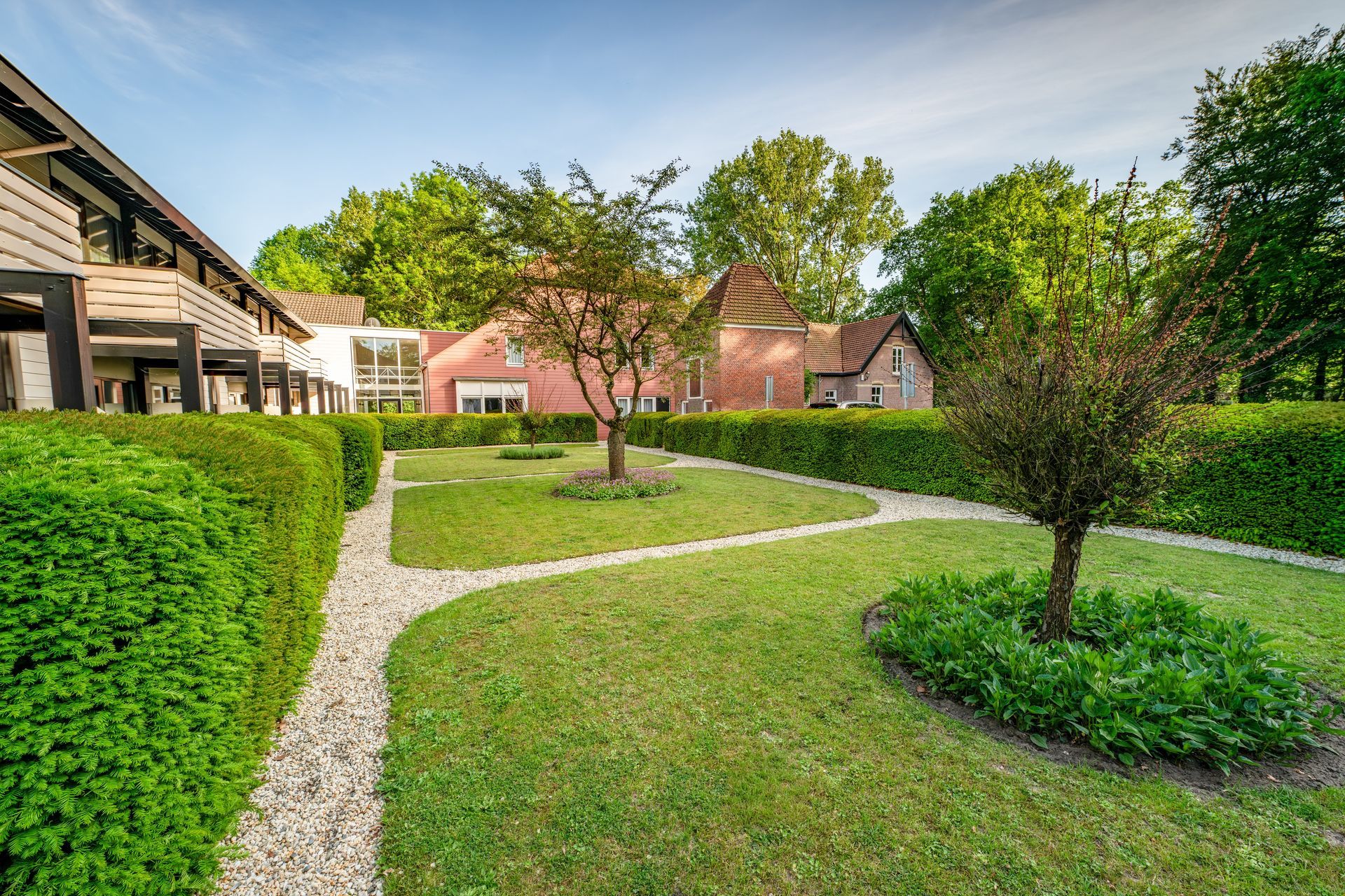 Binnentuin met hagen, grindpaden, een kleine boom en moderne gebouwen onder een helderblauwe hemel.