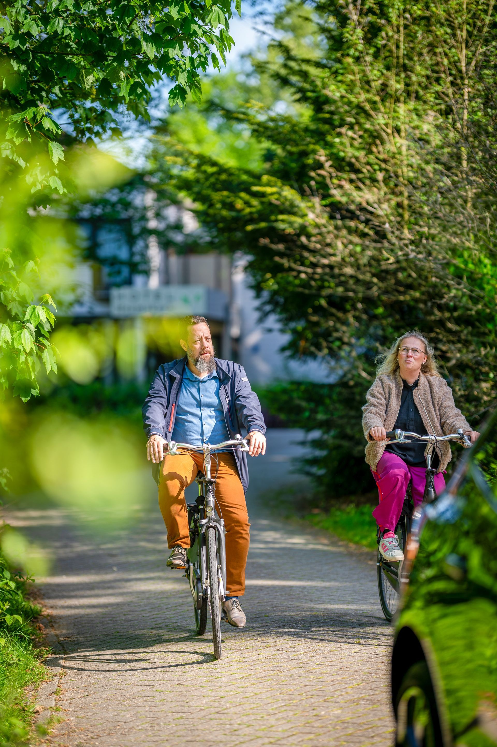 Twee fietsers rijden over een zonnig, met bomen omzoomd pad, de een in een oranje broek en de ander in een magenta broek.