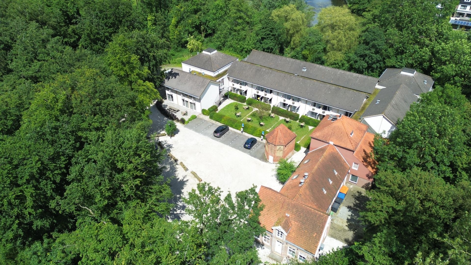 Een luchtfoto van Hotel Heemskerk, omringd door weelderig groen, een parkeerplaats met auto's en een binnenplaats met een gazon.