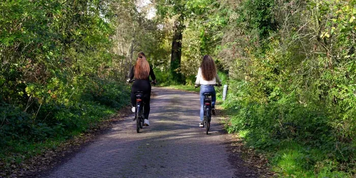 Two people cycling on a paved path through a green, wooded area in Heemskerk. They are riding in the same direction, away from the camera.