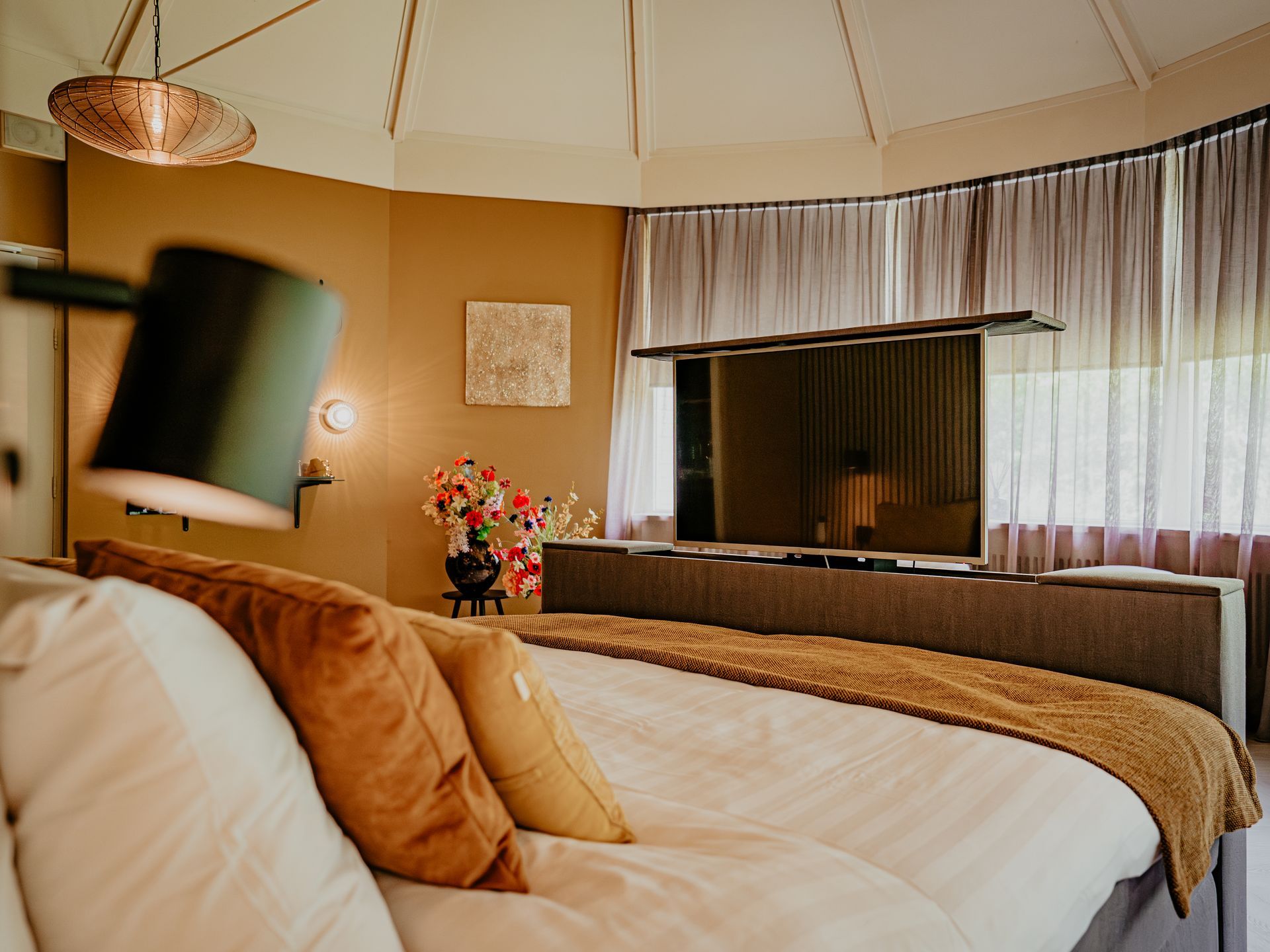 Luxury suite at Hotel Heemskerk with a stylishly made bed with decorative pillows, flowers, and a flat-screen TV rising from the foot of the bed.