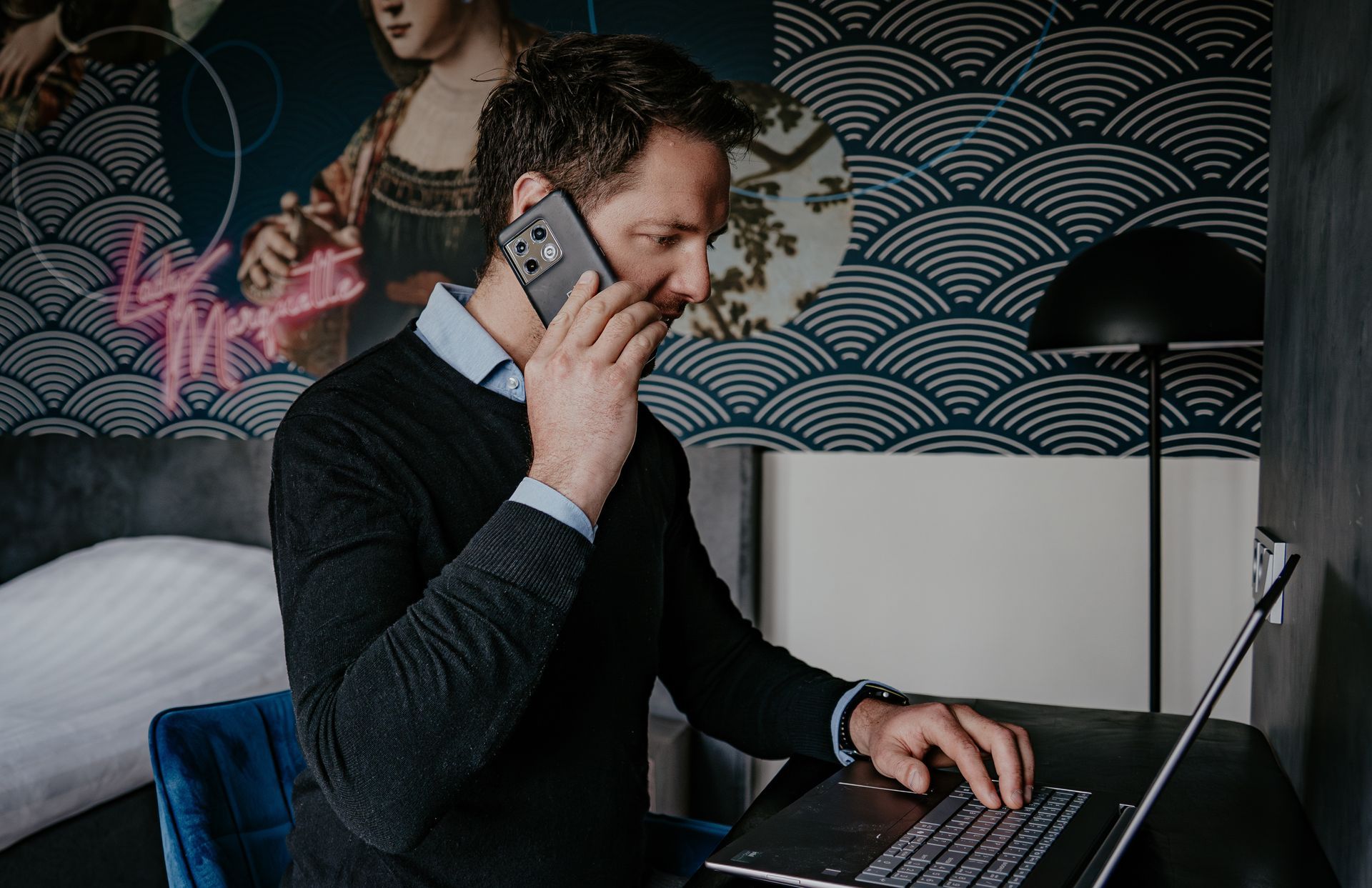 A man working in the single room at Hotel Heemskerk, sitting at the desk with a laptop and phone, with artistic wall decoration in the background.