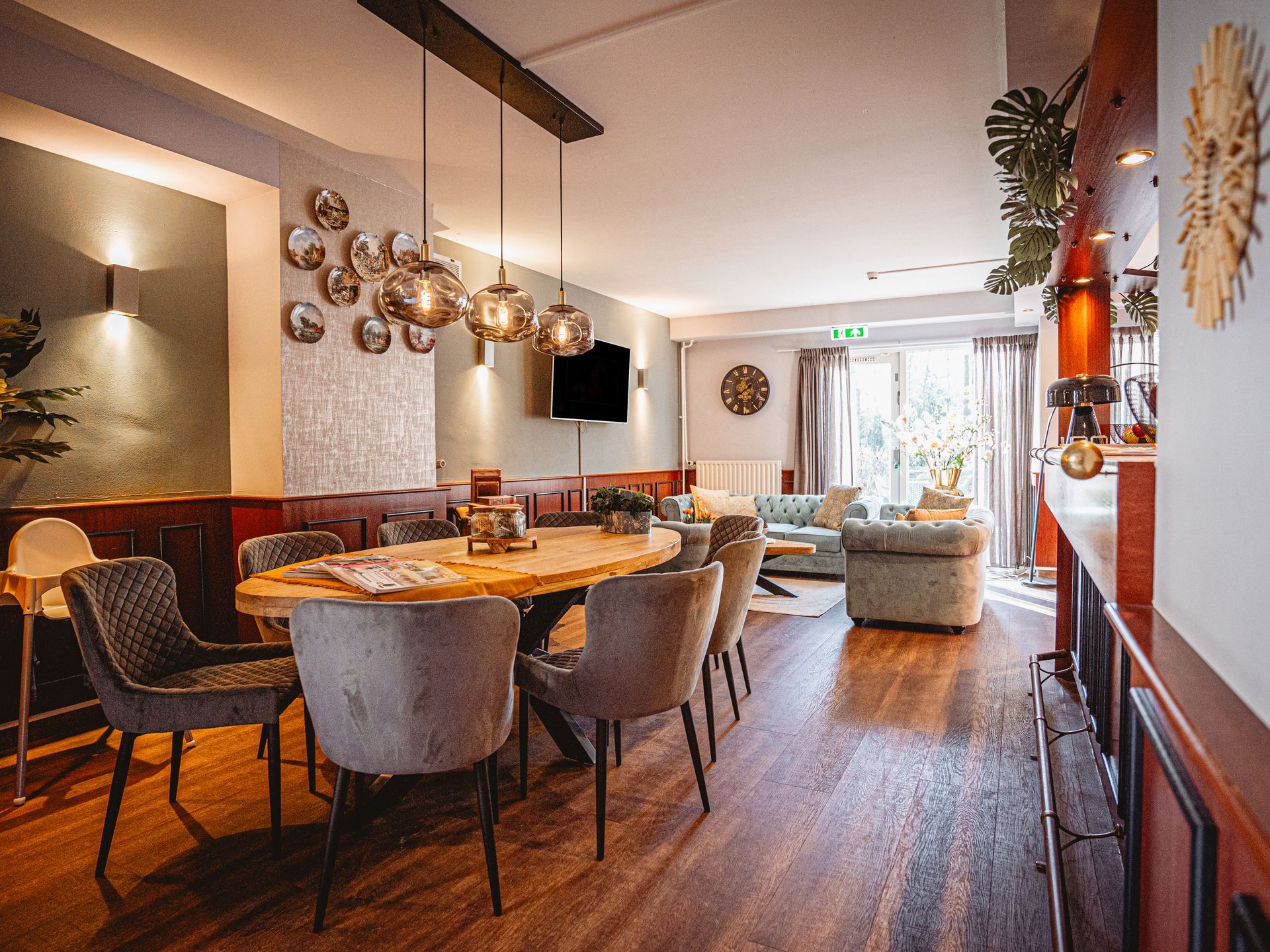Een overzichtsfoto van de lounge van Hotel Heemskerk, met een grote ovale houten tafel omringd door grijze gestoffeerde stoelen, moderne hanglampen erboven, een zithoek met blauwe en grijze banken in de achtergrond, en houten vloeren door de hele ruimte.