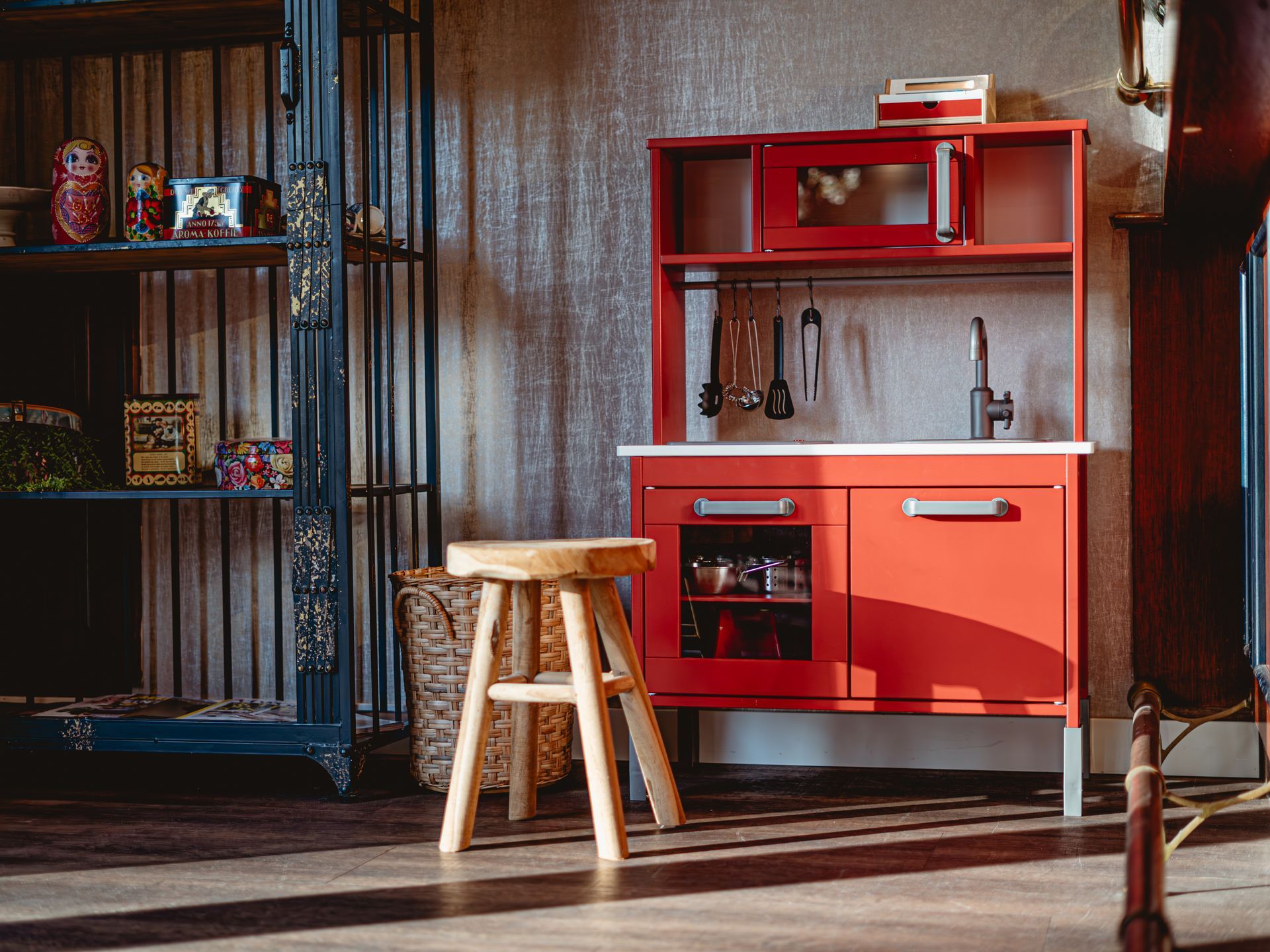 Een speelhoek met een rood speelkeukentje met een oven, magnetron, gootsteen en kookgerei, naast een houten krukje en een donkerblauwe open kast met speelgoed en decoratieve items.