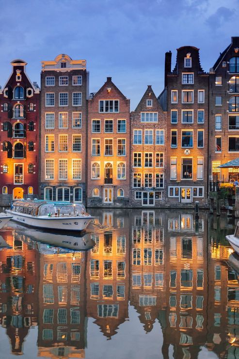Een sfeervolle foto van een Amsterdamse gracht met verlichte, smalle grachtenpanden langs de kade. Een rondvaartboot ligt aangemeerd op het rustige water, waarin de reflecties van de gebouwen en de lichten prachtig zichtbaar zijn onder een blauwe avondlucht.