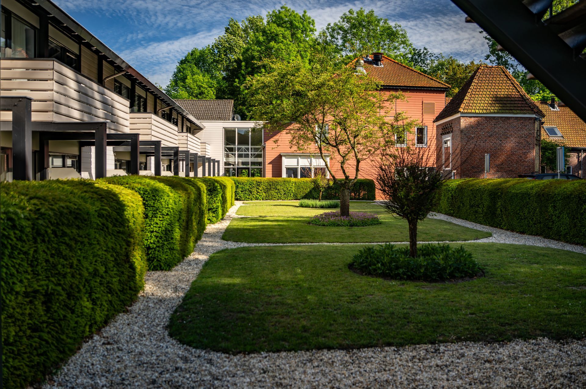 Landscaped courtyard with hedges, gravel paths, and brick and modern buildings under a blue sky