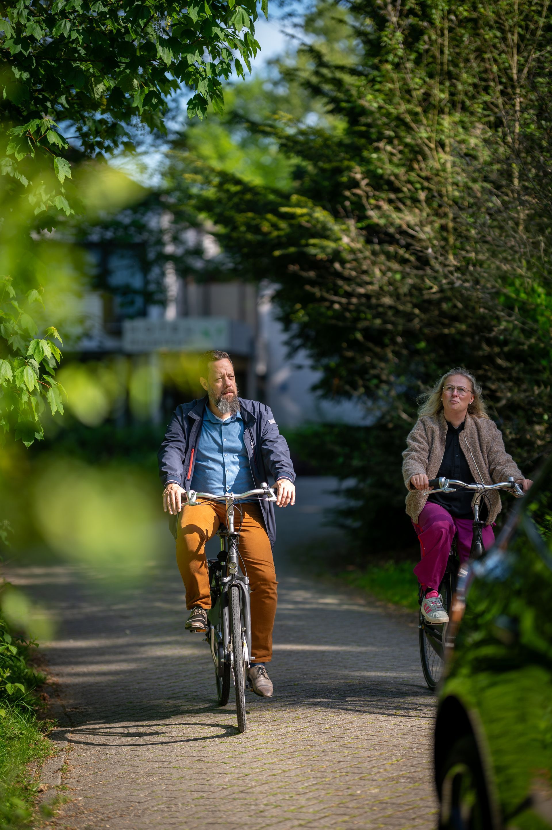 Two cyclists riding on a leafy path, framed by blurred green branches.