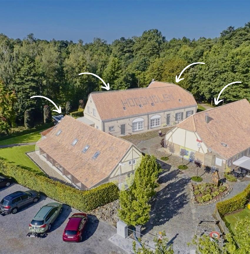 Een luchtfoto van een bakstenen gebouw met drie secties, omgeven door bomen en een parkeerterrein.