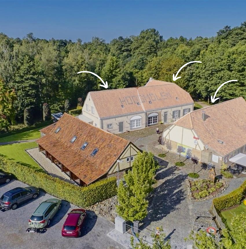 Luchtfoto van diverse gebouwen met rode pannendaken, omgeven door bomen en een parkeerplaats met auto's.