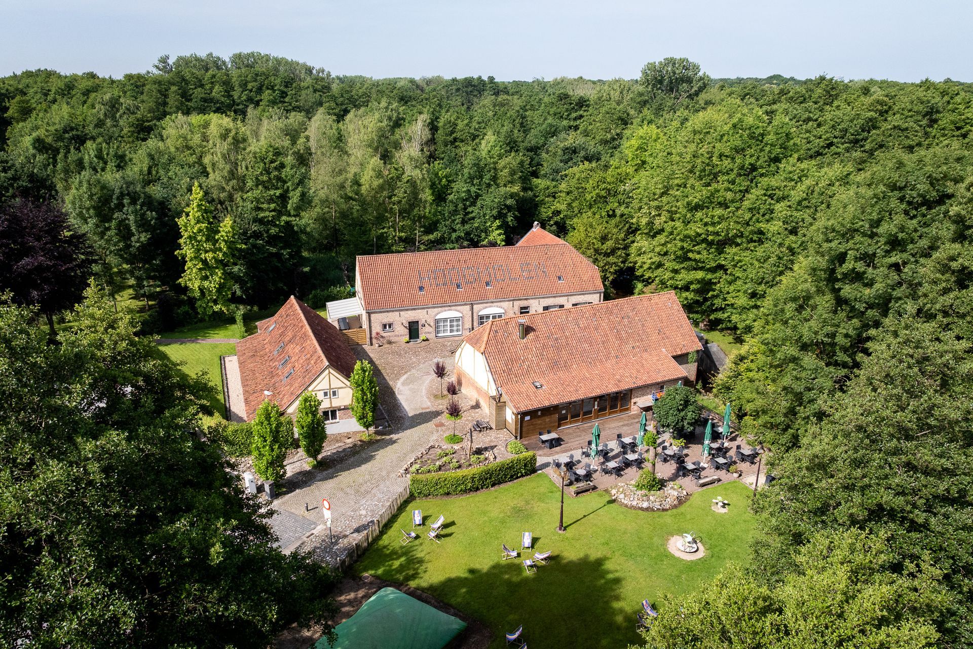 Gebouw met rode pannendaken, omgeven door bomen en een groen gazon. Buiten zitgedeelte zichtbaar.