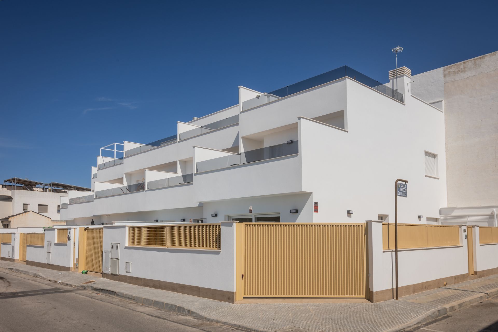 Wit appartementencomplex met balkons, een geel hek en een helderblauwe lucht.