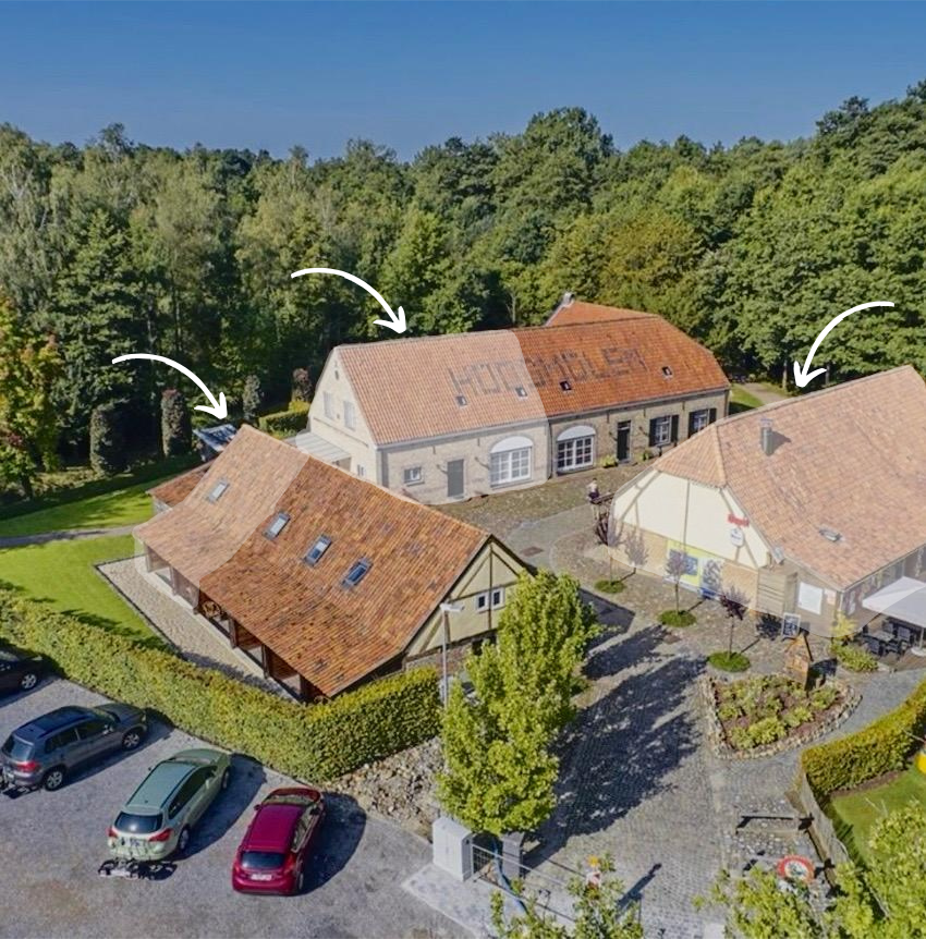 Luchtfoto van een complex van bakstenen gebouwen met rode pannendaken, omgeven door groene bomen en een parkeerplaats.