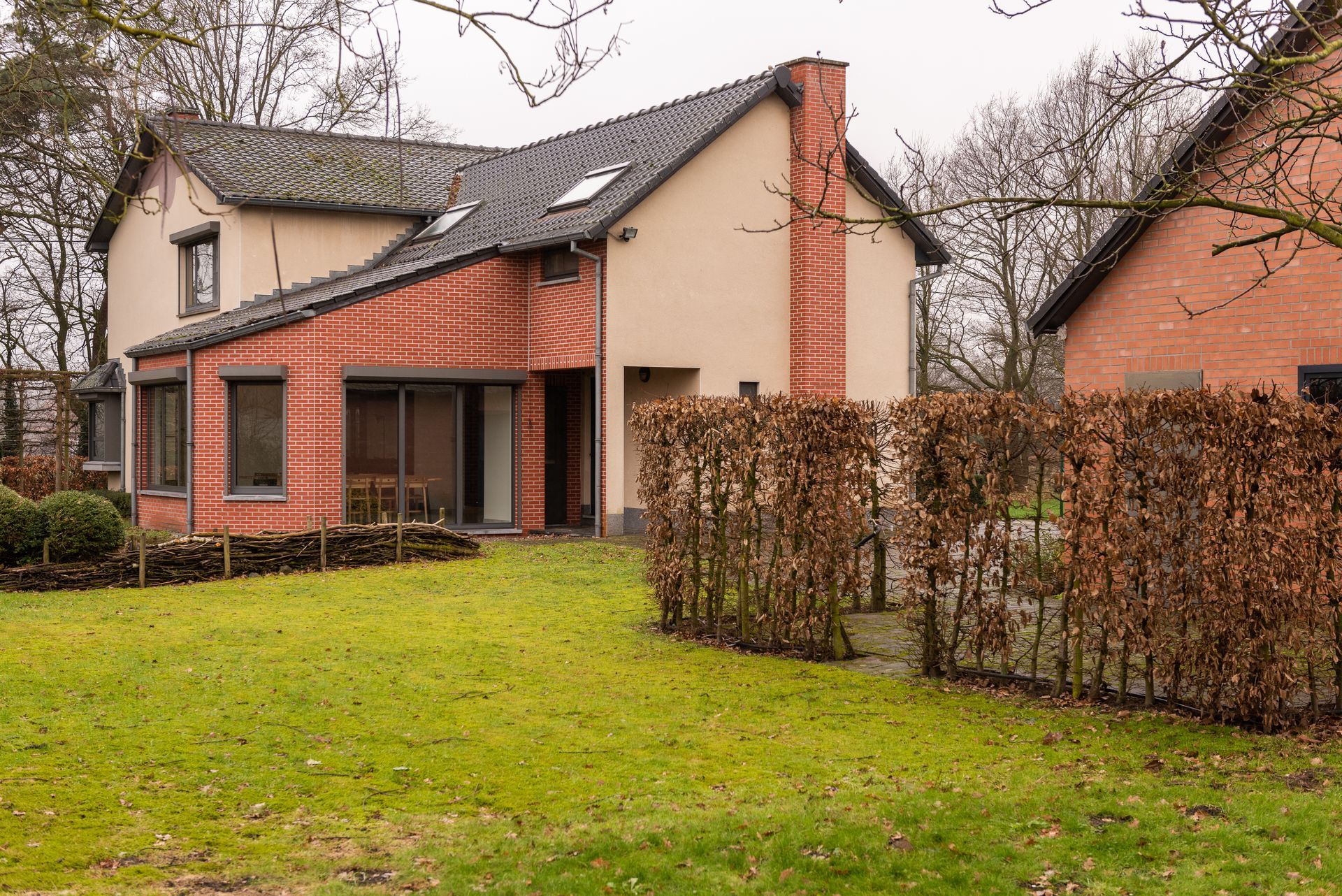 Een huis met twee verdiepingen, een gevel van baksteen en beige accenten, een grasveld en een haag als schutting.