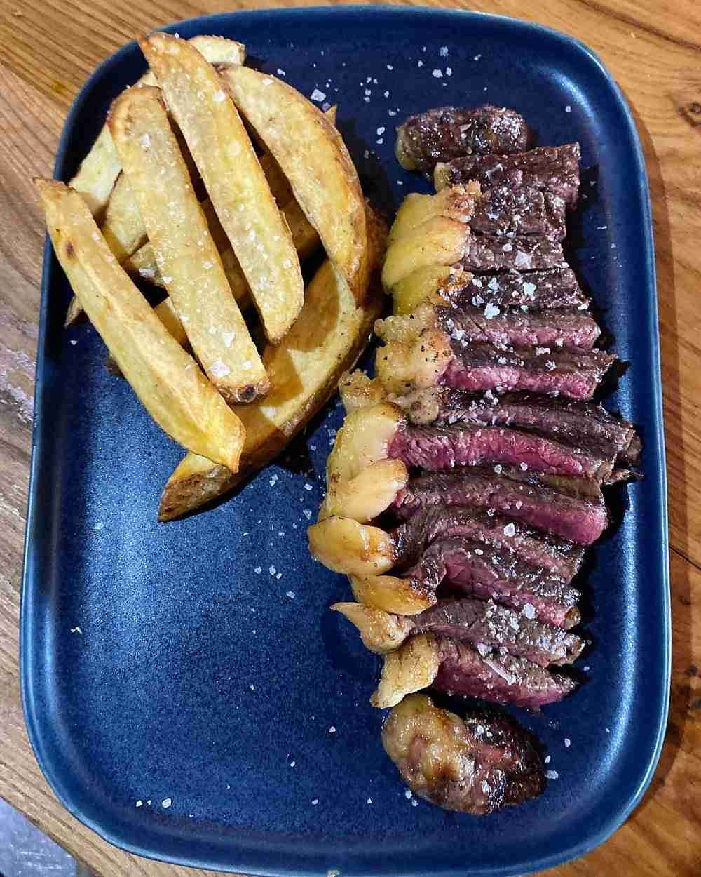 Plato azul con filete en rodajas y patatas fritas gruesas sobre una mesa de madera.