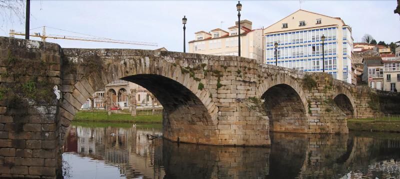 Puente de piedra con aberturas arqueadas sobre un río reflectante; edificios y cielo visibles en el fondo.