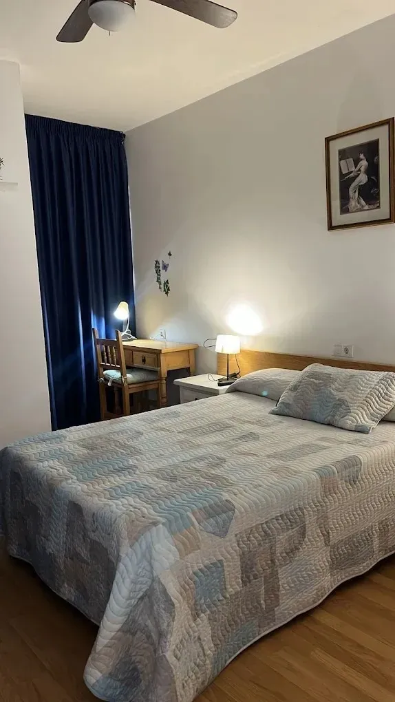 Dormitorio con cama, escritorio, cortinas azules y obras de arte. Ventilador de techo y dos lámparas.