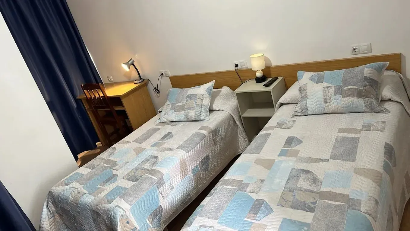 Dos camas individuales en una habitación con escritorio, mesita de noche, lámparas y colchas con estampados azules.