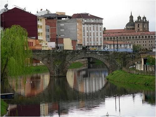 Arcos de puente de piedra sobre un río tranquilo; edificios y una iglesia al fondo.
