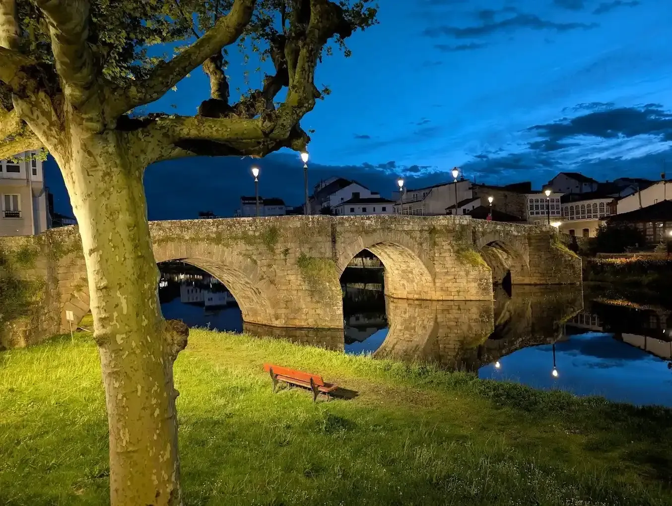 Puente de piedra sobre aguas tranquilas al anochecer con reflejos y edificios iluminados.