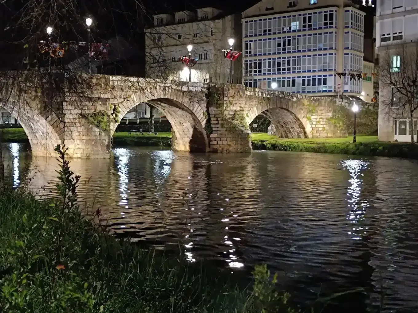 Puente de piedra sobre un río de noche, iluminado por farolas. Edificios al fondo.