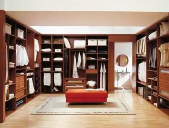 Vestidor con estanterías de madera oscura, ropa blanca y un puf rojo sobre una alfombra clara.