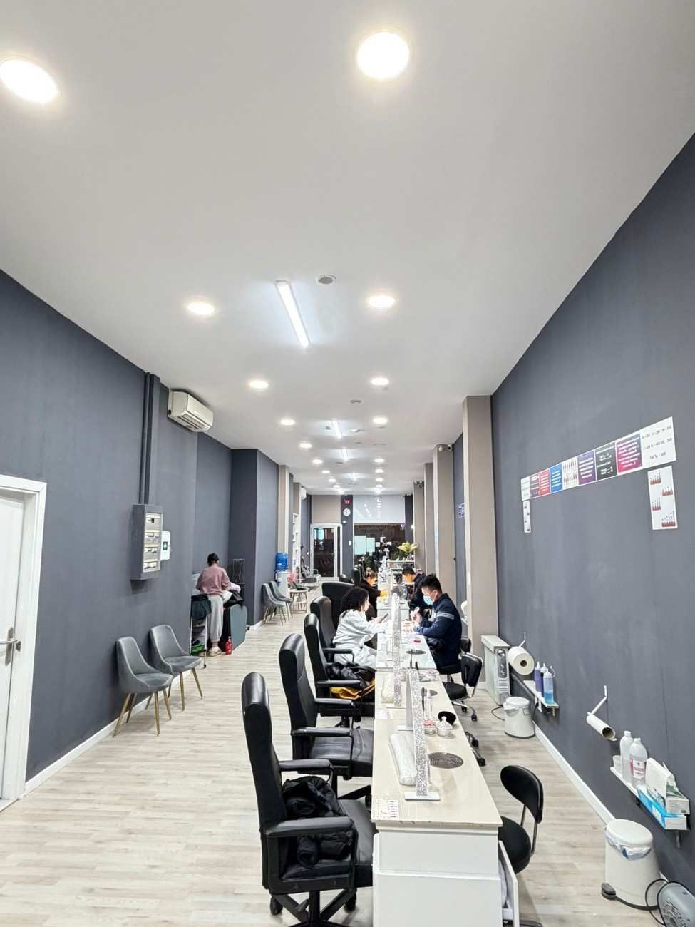 Interior de salón de uñas con paredes grises, mostradores blancos y clientes sentados en estaciones.