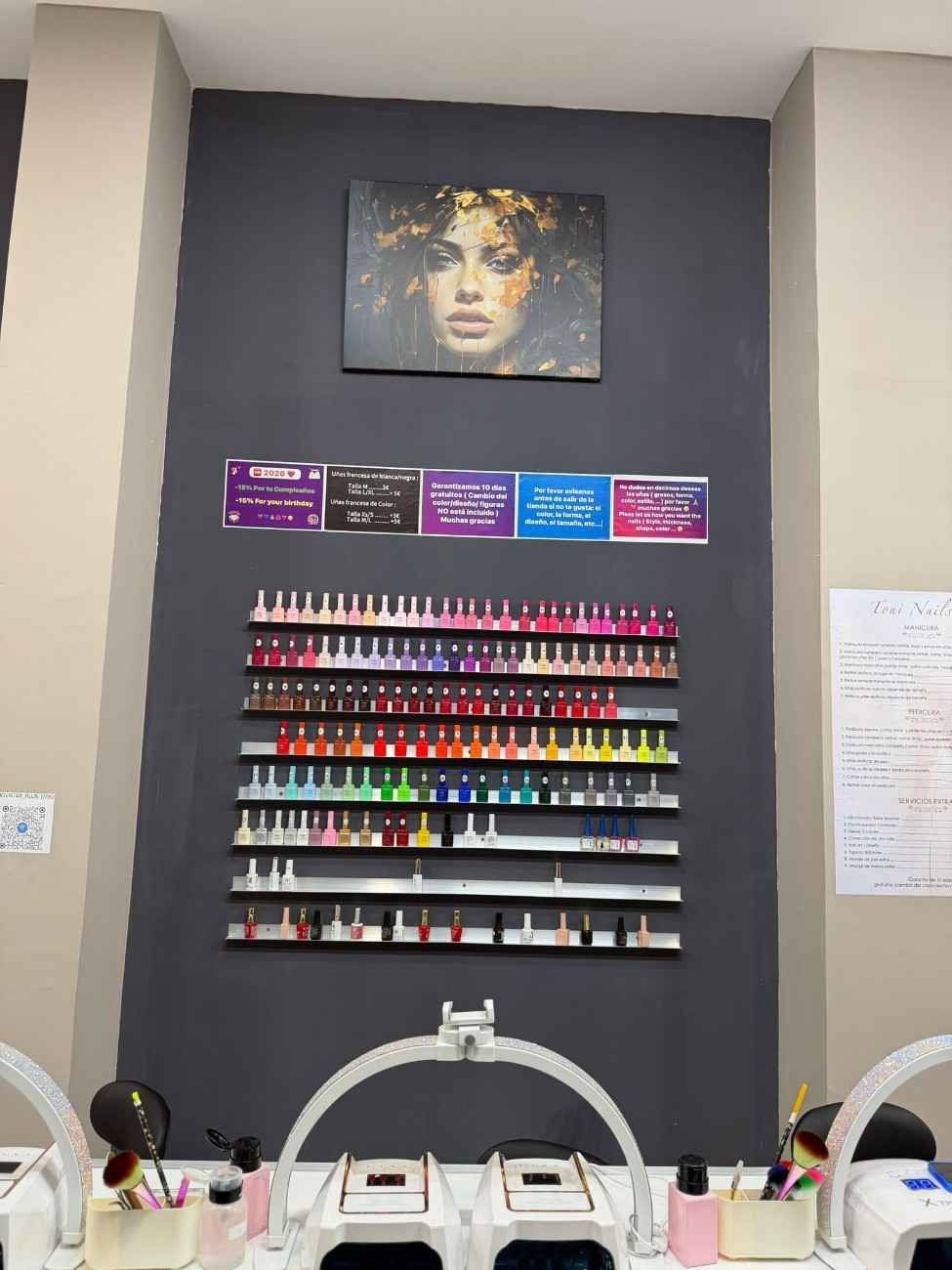 Interior de salón de uñas con filas de colores de esmaltes de uñas exhibidos en una pared gris debajo de un retrato.