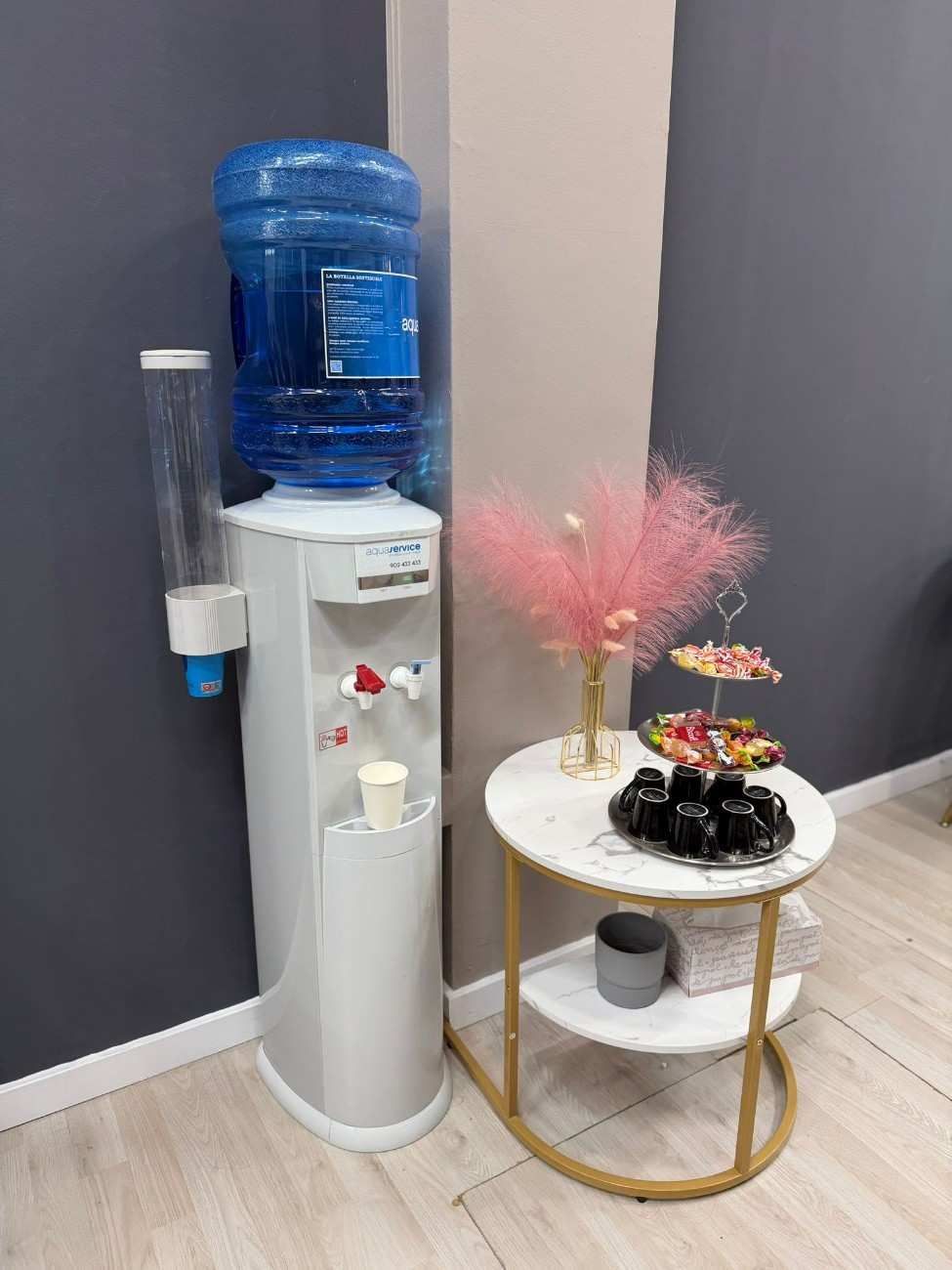 Enfriador de agua blanco al lado de una pequeña mesa con golosinas y un arreglo floral decorativo de color rosa.