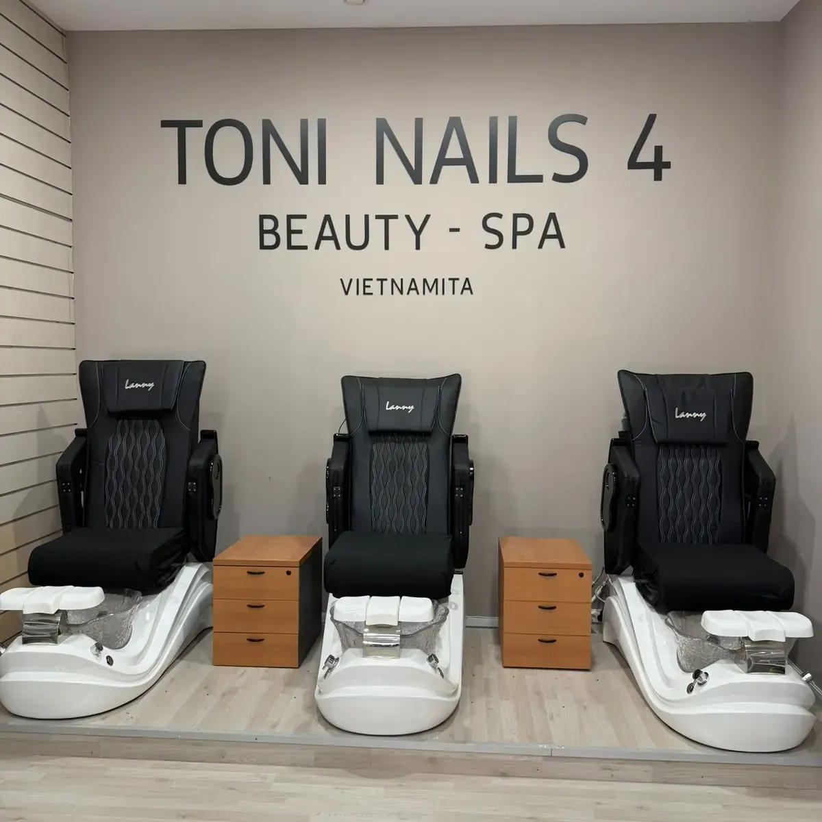 Toni Nails 4: tres sillas de pedicura negras, pared beige con cartel, pequeños cajones de madera.
