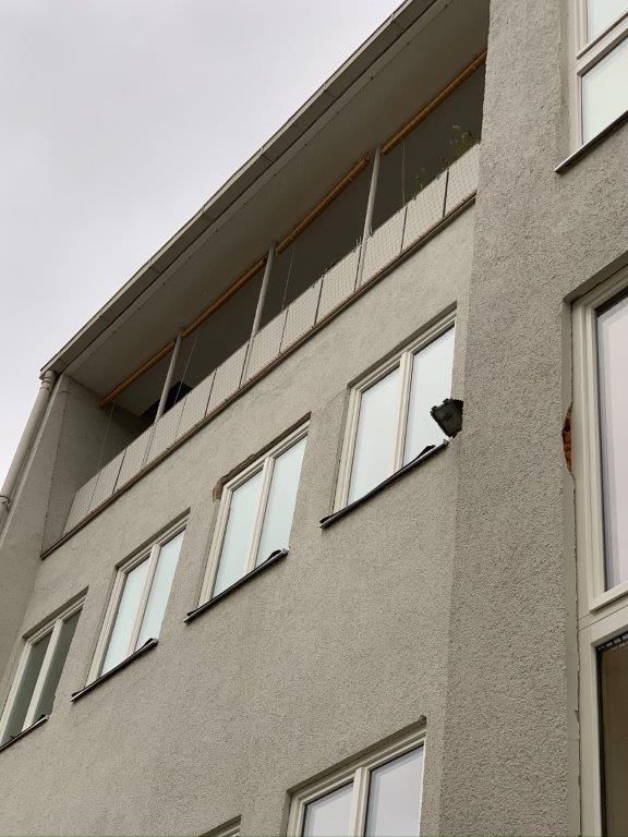 Graues Gebäude mit mehreren weiß gerahmten Fenstern und einem Balkon mit Holzrahmen unter einem bewölkten Himmel.