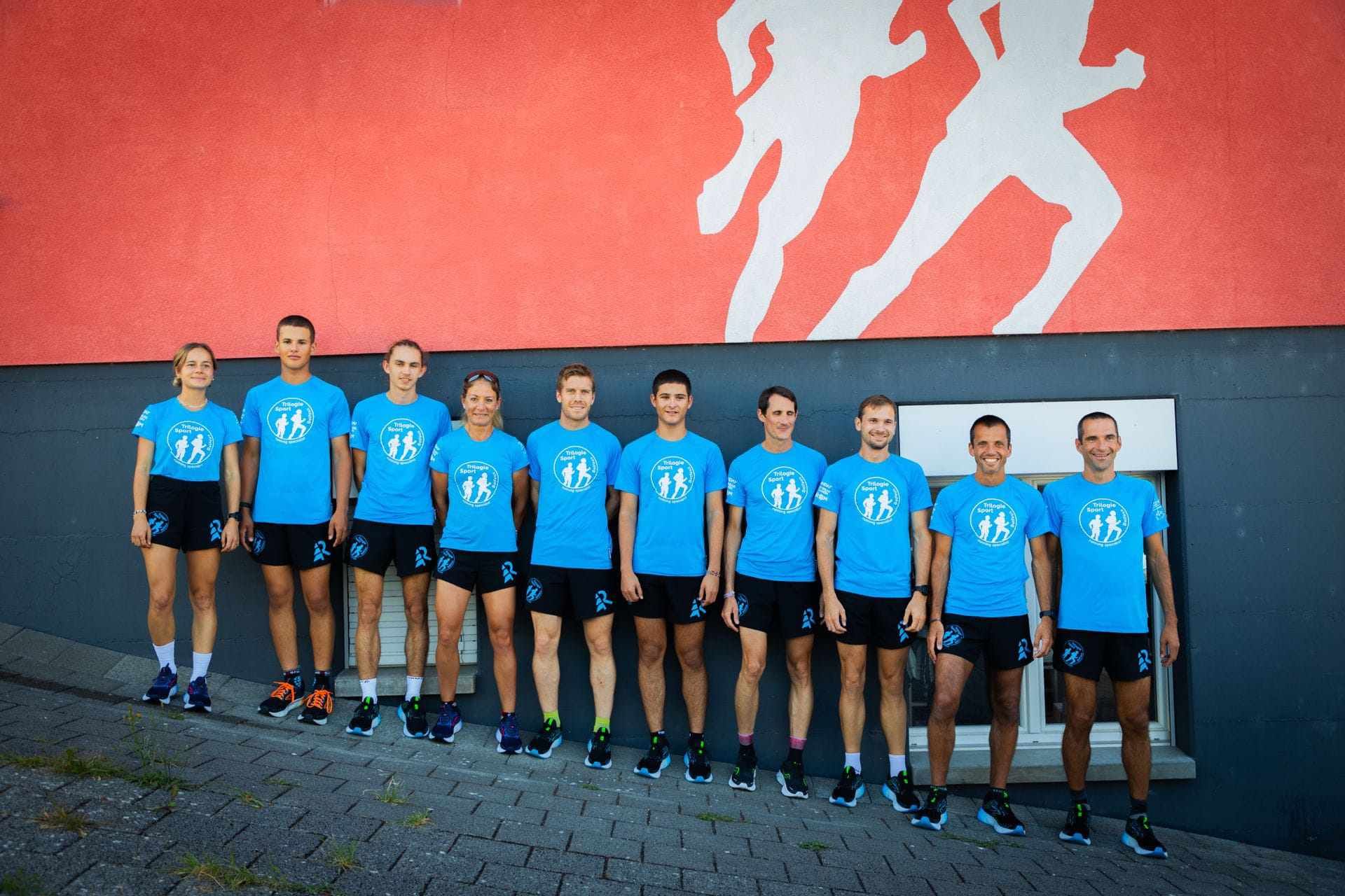 Un groupe de coureurs en maillots bleus et shorts noirs se tient devant un mur rouge orné de silhouettes de coureurs.