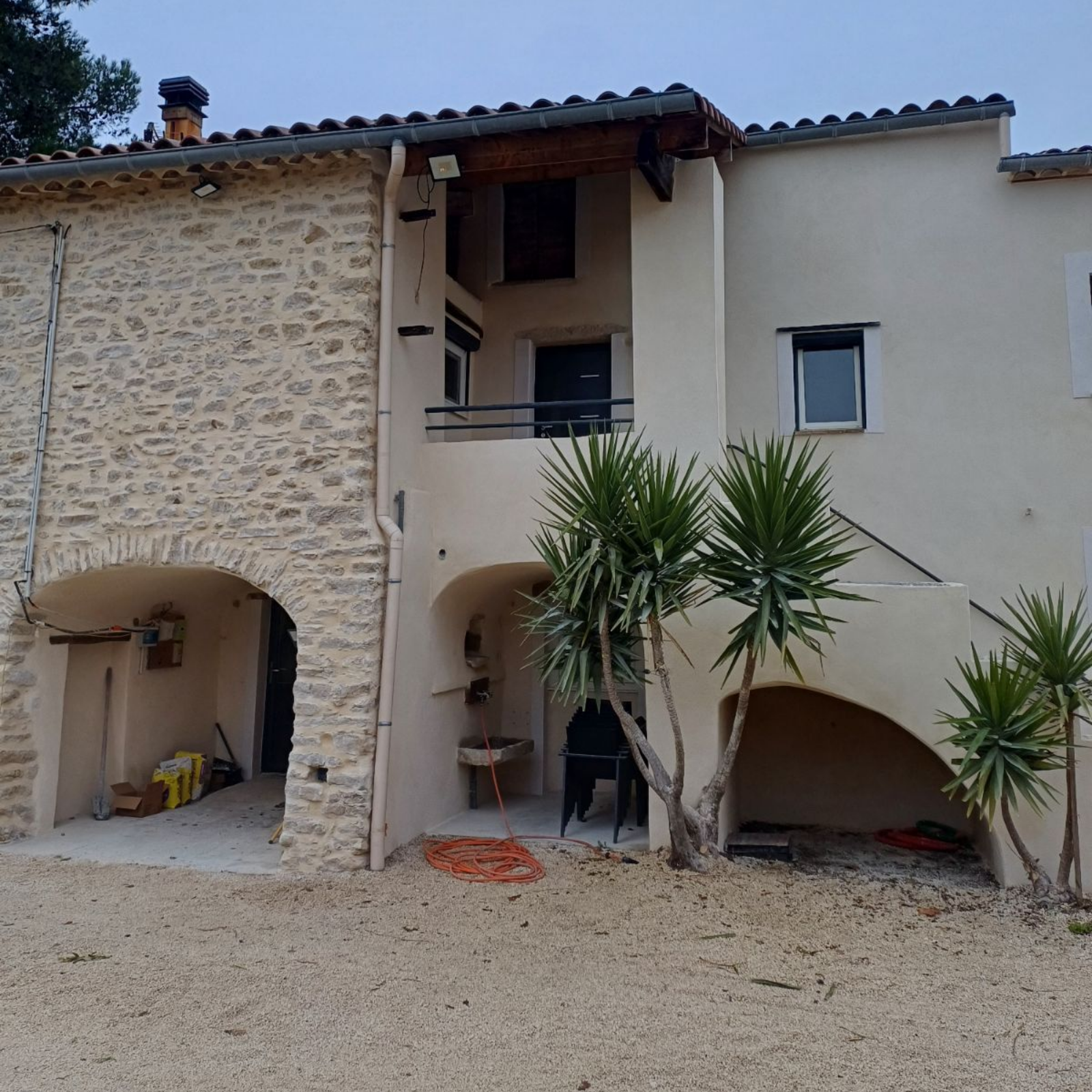 Maison avec toit de tuiles rouges, murs blancs, échafaudage et clôture blanche devant une cour