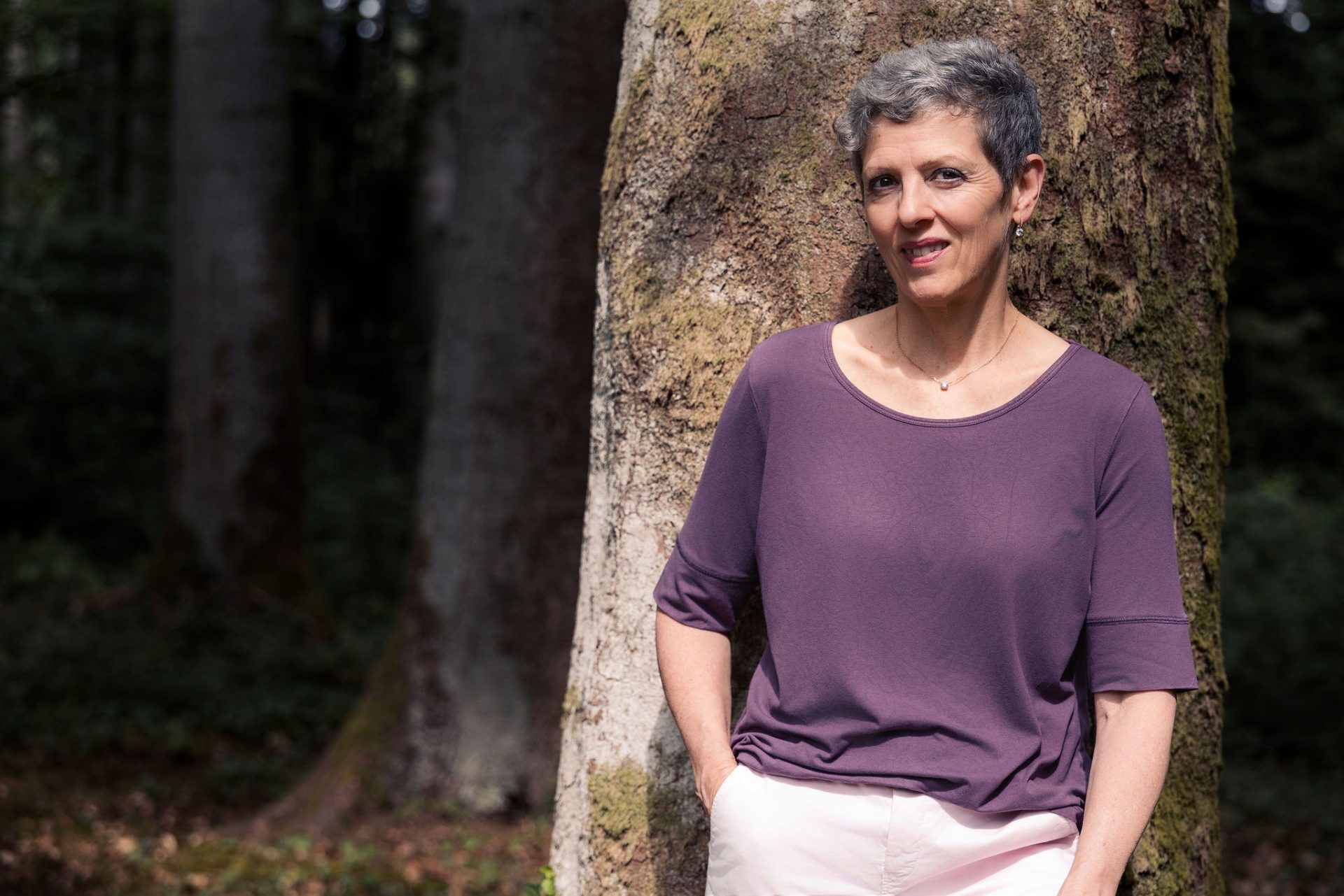 Frau in einem lila Oberteil lehnt an einem Baum in einem sonnenbeschienenen Wald.