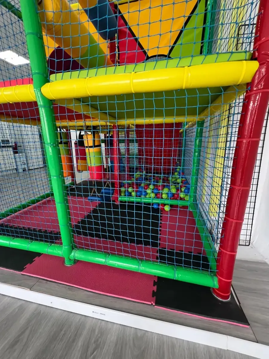 Estructura de juego para interiores con soportes verdes y rojos, red de colores, piscina de pelotas y suelo acolchado.