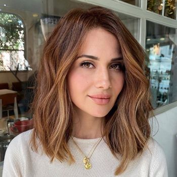 Una mujer con cabello largo y castaño lleva un suéter blanco y un collar de oro.