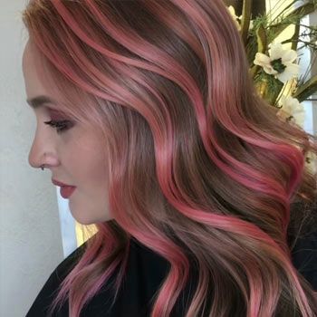 Una mujer con cabello largo castaño con reflejos rosados está parada frente a un jarrón de flores.