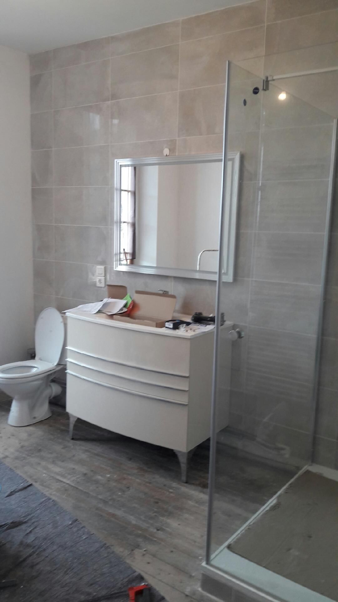 Salle de bains avec meuble blanc, toilettes et douche en verre, murs carrelés gris et sol imitation bois